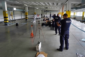 Widok na umundurowanych policjantów i funkcjonariuszy ubranych w białe kombinezony przeprowadzających oględziny na krytym policyjnym parkingu