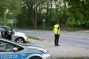 Policjant stoi przy drodze, w ręku trzyma tarczę do zatrzymywania pojazdów