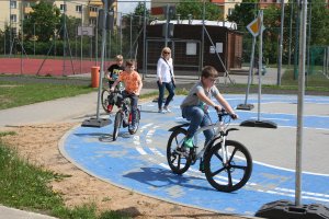 Troje dzieci jadących na rowerach na miasteczku ruchu drogowego, w tle kontrolująca nauczycielka