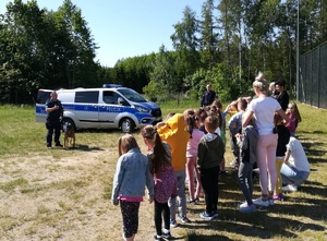 policjant z psem sluzbowym stoi przy dzieciach, w tle radiowóz