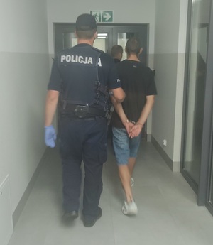 policjant prowadzi zatrzymanego, który na rękach ma załozone kajdanki