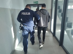 policjant prowadzi zatrzymanego mężczyzne, który ma założone kajdanki na nogi