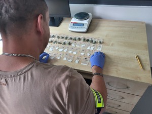 policjant siedzi przy biurku, na którym jest elektroniczna waga oraz woreczki stunowe z zawartościa suszu roslinnego i biała substacją, woreczki sa ponumerowane od 1 do 49
