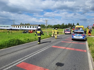 uszkodzony samochód, pociag oraz strazany, na drodze stoi samochód policji, karetka pogotowie oraz samochody strażackie