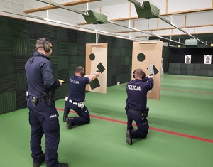 dwóch policjantów w pozycji kleczacej oddaje strzały zza przesłony z trzema otworami