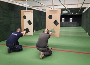 dwóch policjantów w pozycji kleczacej oddaje strzały zza przesłony z trzema otworami