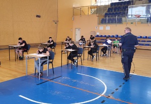 zawodnicy siedzą przy stolikach i rozwiązują test, policjant stoi przy uczniach