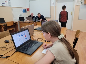 dziewczynka siedzi przed ekranem laptopa i rowiązuje test, z tyłu dwóch policjantówsiedzi w ławce i sparwdzają testy