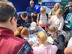 dzieci przy stoliku rozwiązują krzyzówke, a obok stoi policjant i obserwuje