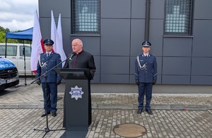na mównicy stoi Ksiądz kanonik, po obu stronach stoja policjanci z tyłu stoi stojak z flagami rzeczypospolitej Polskiej