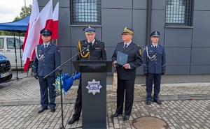 na mównicy stoi z-ca Komendanta Straży Pożarnej, obok Komendant Strazy Miejskiej, po obu stronach stoja policjanci z tyłu stoi stojak z flagami rzeczypospolitej Polskiej