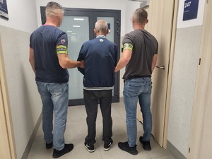dwóch policjantów w umundurowaniu cywilnym z założonymi opaska na ręku z napisem policja trzyma za recę mężczyznę. Wszyscy odwróceni sa plecami.