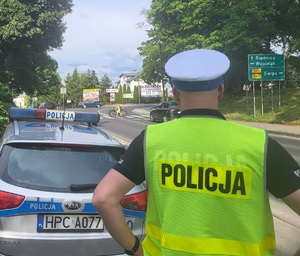 policjant stoi przy radiowozie i obserwuje przejście dla pieszych gdzie przechodza osoby z rowerami