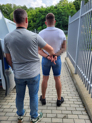 policjant wyprowadza zatrzymanego mężczyznę, który ma rękach załozone kajdanki