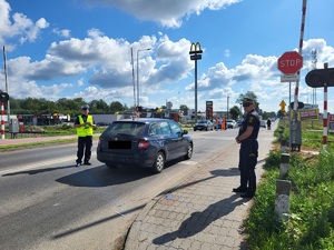 policjant oraz strażnik miejski stoją na drodze, gdzie przez przejazd kolejowy przejeżdża samochód