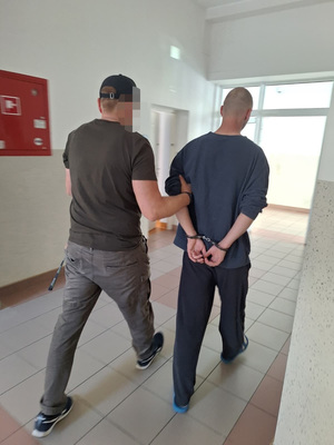 policjant w ubiorze cywilnym prowadzi zatrzymanego męzczyzne, który ma załozone kajdanki na rece trzymane z tyłu