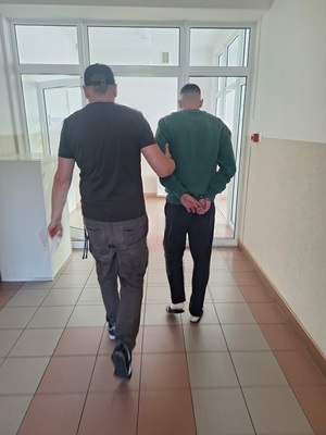 policjant w ubiorze cywilnym prowadzi zatrzymanego męzczyzne, który ma załozone kajdanki na rece trzymane z tyłu