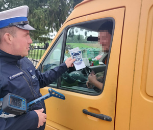 policjant stoi przy samochodzie i wrecza kierowcy naklejkę z napisem &quot;mistrzu zwolnij&quot;