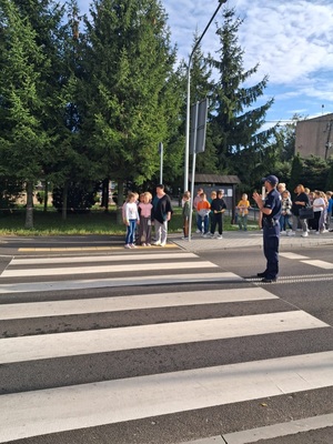 policjant stoi przed przejsciem dla pieszych na drodze, na chodniku stoja dzieci