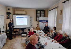 policjantka stojąca przy ekranie prowadzaca prelekcję, przy stole siedza uczestnicy debaty