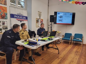 dwóch policjantów oraz strażąk siedza przy stole, na ekranie telewizora wyświetlana jest prezentacja