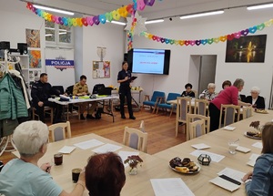 policjantka stoi i prezentuje prelekcję, na stołem siedzi policjant i strażak, po drugiej stronie sali przy stołach siedza uczestnicy spotkania