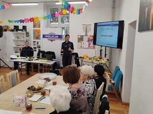 policjantka stoi przy ekranie i prowadzi prelekcję, przy stołach siedzauczestnicy spotkania