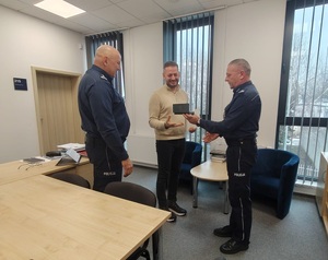 trzy osoby na zdjęciu, komendant wręcza upominek jubilatowi