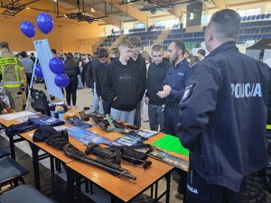 policyjne stanowisko przy którym stoi kilka osób, na stoliku widoczny jest sprzet w postaci jednostek broni, ale także ulotki, gazety policyjne, gażdzety reklamowe