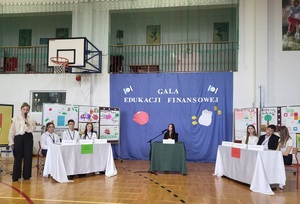 scena, na ktorej przy stolikach siedzą dwie drużyny oznaczone jako drużyna inwestycji i drużyna oszczedzania