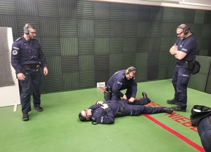 policjant udziela pomocy drugiemu policjantowi, który leży na ziemi, obok stoi dwóch policjantów prowadzacych strzelanie