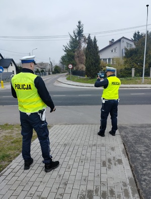 dwóch policjantów stojących przy drodze, jeden z nich trzyma miernik predkosci