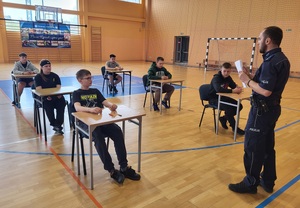 sześciu zawodników siedzi w ławkach, a przed nimi stoi policjant