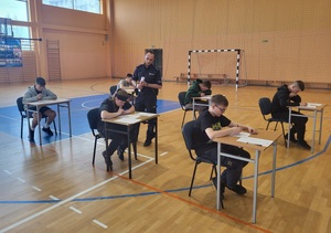 zawodnicy rozwiązują testy, między ławkami chodzi policjant