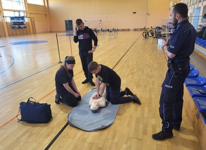 trzyobowa druzyna udziela pomocy przedmedycznej przy użyciu fantoma, obok stoi policjant