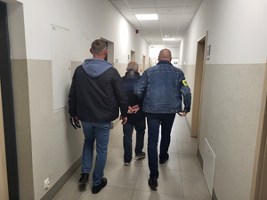 dwóch policjantów prowadzi zatrzymanego mężczyznę, który ma załozone kajdanki przez korytarz