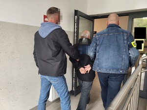 dwóch policjantów prowadzi zatrzymanego mężczyznę, który ma załozone kajdanki przez korytarz
