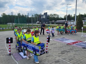 dzieci przebrane  w samochodziki poruszaja sie po miasteczku ruchu drogowego, a między nimi porusza się policjant, który wskazuje jak prawidłowo poruszac się po drodze