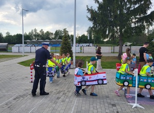 dzieci przebrane  w samochodziki poruszaja sie po miasteczku ruchu drogowego, a między nimi porusza się policjant, który wskazuje jak prawidłowo poruszac się po drodze