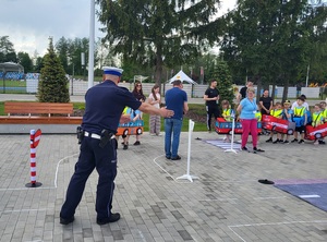 dzieci przebrane  w samochodziki poruszaja sie po miasteczku ruchu drogowego, a między nimi porusza się policjant, który wskazuje jak prawidłowo poruszac się po drodze