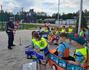 dzieci przebrane  w samochodziki poruszaja sie po miasteczku ruchu drogowego, a między nimi porusza się policjant, który wskazuje jak prawidłowo poruszac się po drodze