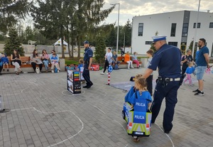 dzieci przebrane  w samochodziki poruszaja sie po miasteczku ruchu drogowego, a między nimi porusza się policjant, który wskazuje jak prawidłowo poruszac się po drodze