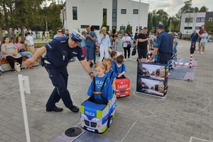 dzieci przebrane  w samochodziki poruszaja sie po miasteczku ruchu drogowego, a między nimi porusza się policjant, który wskazuje jak prawidłowo poruszac się po drodze