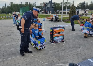 dzieci przebrane  w samochodziki poruszaja sie po miasteczku ruchu drogowego, a między nimi porusza się policjant, który wskazuje jak prawidłowo poruszac się po drodze