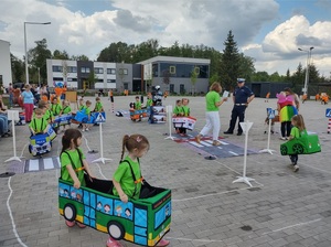 dzieci przebrane  w samochodziki poruszaja sie po miasteczku ruchu drogowego, a między nimi porusza się policjant, który wskazuje jak prawidłowo poruszac się po drodze