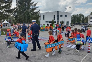dzieci przebrane  w samochodziki poruszaja sie po miasteczku ruchu drogowego, a między nimi porusza się policjant, który wskazuje jak prawidłowo poruszac się po drodze