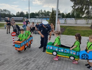 dzieci przebrane  w samochodziki poruszaja sie po miasteczku ruchu drogowego, a między nimi porusza się policjant, który wskazuje jak prawidłowo poruszac się po drodze