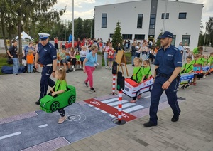 dzieci przebrane  w samochodziki poruszaja sie po miasteczku ruchu drogowego, a między nimi porusza się policjant, który wskazuje jak prawidłowo poruszac się po drodze
