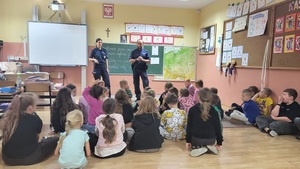 policjanci stoja przed dziecmi w klasie, które siedza na dywanie i słuchaja prelekcji