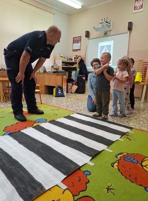dzieci ustawily sie w kolejnce do przejścia dla pieszych, policjant stoi obok i obserwuje zachowanie dzieci